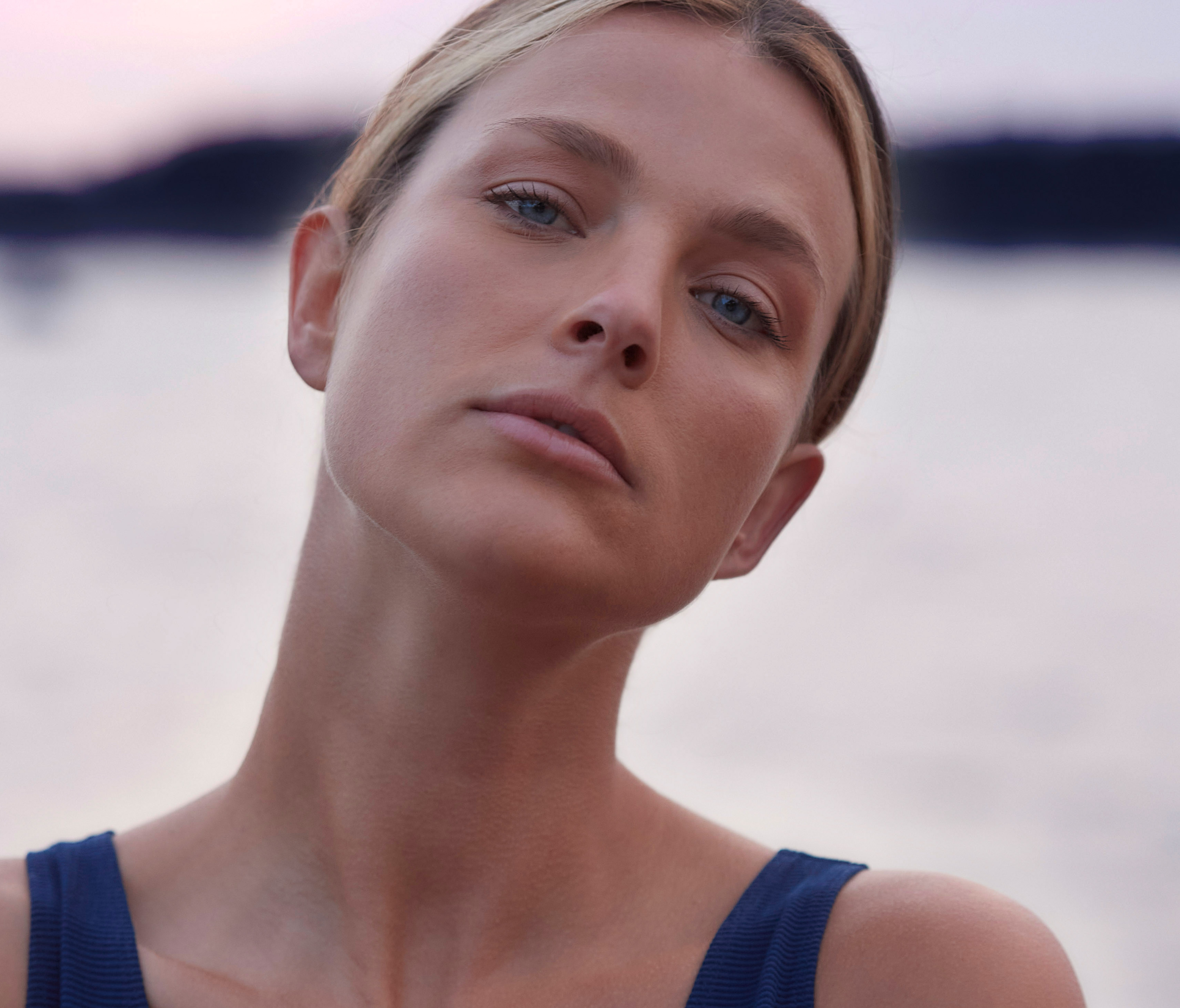A woman at the beach during sunset with her head tilted to the side, gazing at the camera.