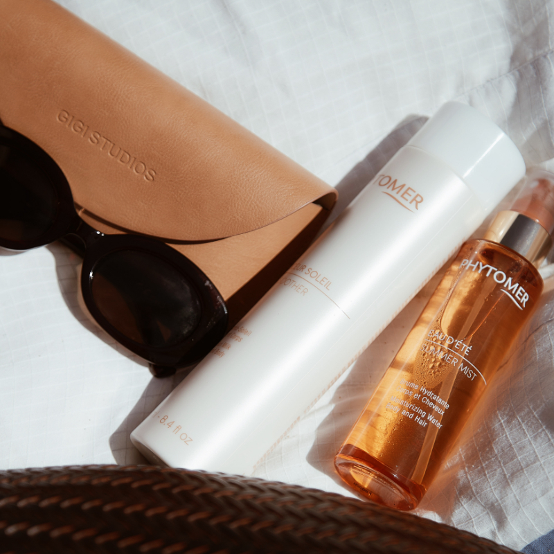 A pair of sunglasses lay next to a Phytomer Sun Soother After-Sun Milk Face and Body Lotion and the Phytomer Summer Mist Moisturizing Water Body & Hair 
