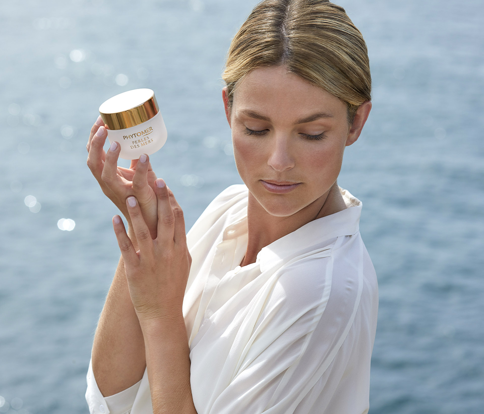 Woman in white clothing holding Phytomer Perles des Mers cream jar against ocean background
