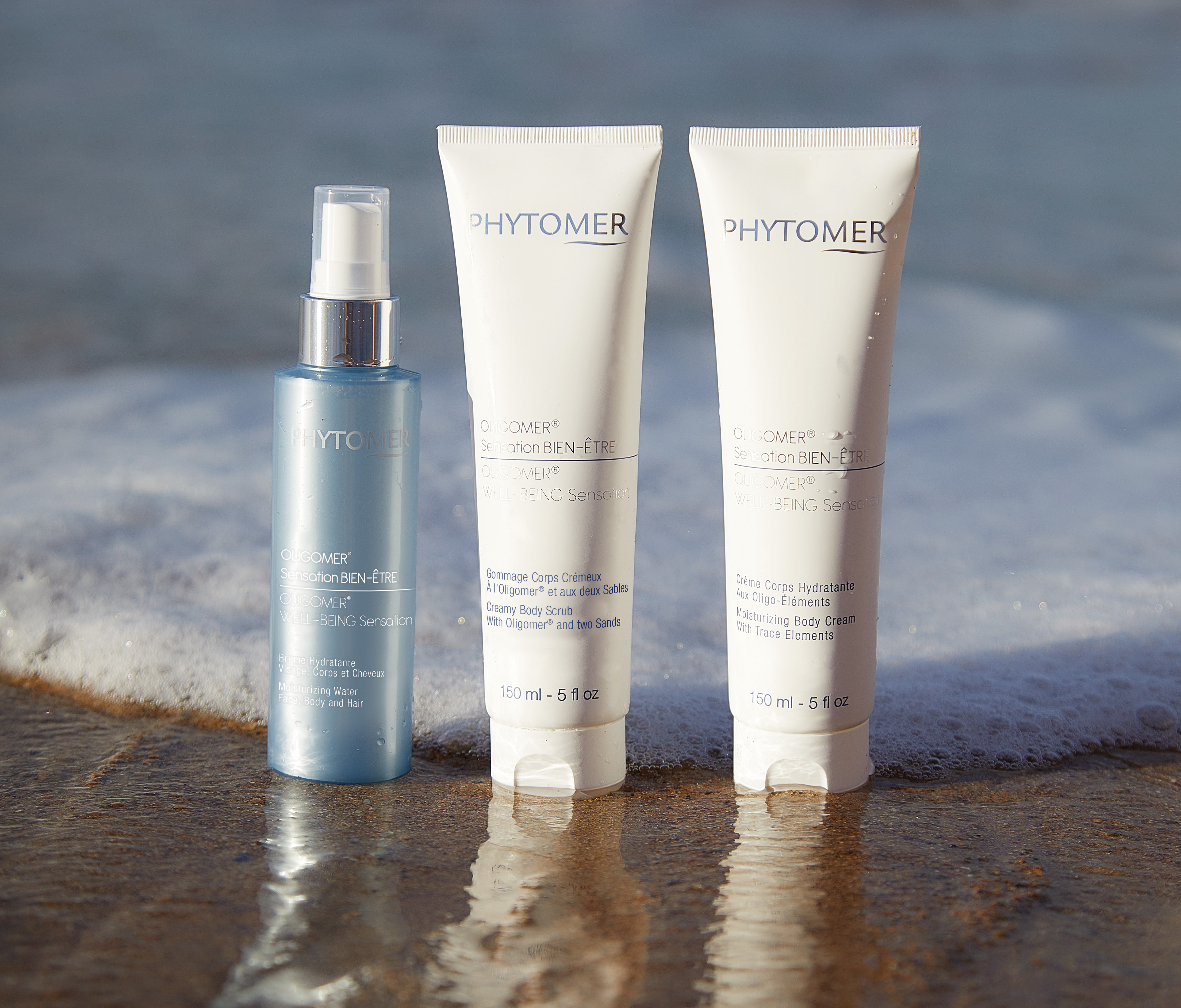 The Oligomer Moisturizing Water face, Body, and Hair, new Oligomer Well-Being Sensation Creamy Body Scrub with Oligomer and two sands, and Oligomer Well-Being Sensation Moisturizing Body Cream with Trace Elements sit side by side on the beach while waves crash around them. 