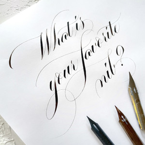 "What is your favorite nib" written in black ink in a pointed calligraphic script with one gold, one blue, and one bronze nib.