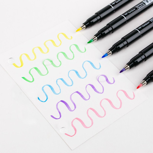 Six pastel Tombow Fudenosuke brush pens uncapped with a wave-like swatch corresponding to each color drawn onto a sheet of paper underneath.