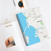 A spiral notebook with a personalized map cover sitting askew on a white desk with papers and sunglasses nearby