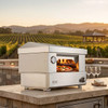 The Outdoor Plus 30 Inch Countertop Videl USA Pizza Oven, outdoor pizza oven with a stainless steel exterior baking a pizza, set on a stone countertop with wine glasses nearby, overlooking a scenic vineyard landscape at sunset.