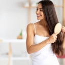 A smiling woman with long shiny healthy brown hair brushing it with a wooden hair brush in a bright bathroom