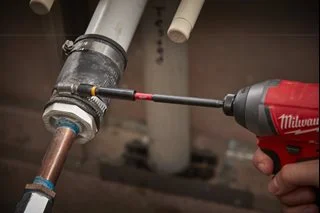 Milwaukee power tool being used in an automotive workshop, with the tool positioned against a vehicle component during a repair or maintenance task. The image demonstrates the product in a practical workshop application.