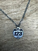 A 15mm number plate shaped titanium pendant hangs from a linked style stainless steel necklace chain. The pendant has the word name to show where it can be personalized with your name and the numbers 1,2,3 to show where it can be personalized with your race number. It is a close-up picture of the necklace sitting on a wooden desk.