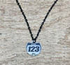 A 15mm number plate shaped titanium pendant hangs from a black linked style stainless steel necklace chain. The pendant has the word name to show where it can be personalized with your name and the numbers 1,2,3 to show where it can be personalized with your race number. It is a close-up picture of the necklace sitting on a wooden desk.