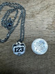 A 15mm number plate shaped titanium pendant hangs from a black linked style stainless steel necklace chain. It is placed next to a U.S. Dime to show a size comparison. It is slightly smaller than the dime. The pendant has the word name to show where it can be personalized with your name and the numbers 1,2,3 to show where it can be personalized with your race number. It is a close-up picture of the necklace sitting on a wooden desk.