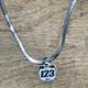 A 15mm number plate shaped titanium pendant hangs from a herringbone style stainless steel necklace chain. The pendant has the word name to show where it can be personalized with your name and the numbers 1,2,3 to show where it can be personalized with your race number. It is a close-up picture of the necklace sitting on a wooden desk.