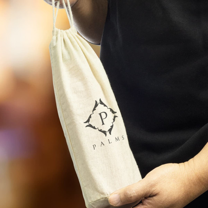 A drink bottle gift bag in natural cotton, featuring a logo, held by a person against a blurred background.