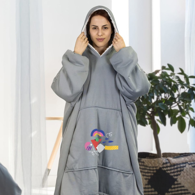 A grey hoodie blanket with a large hood, featuring a logo, worn by a woman in a bright room with plants.