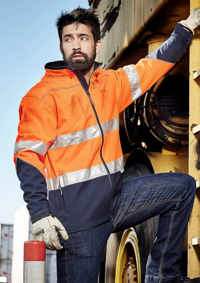 A unisex hi-vis softshell jacket in orange and navy, featuring reflective strips, worn by a person leaning against machinery.