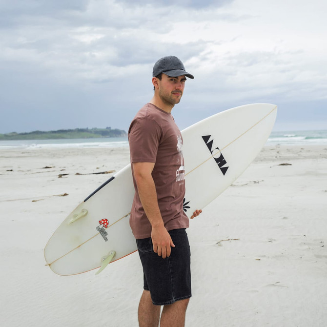 A man stands on a beach holding a surfboard, wearing a brown T-shirt and dark shorts.