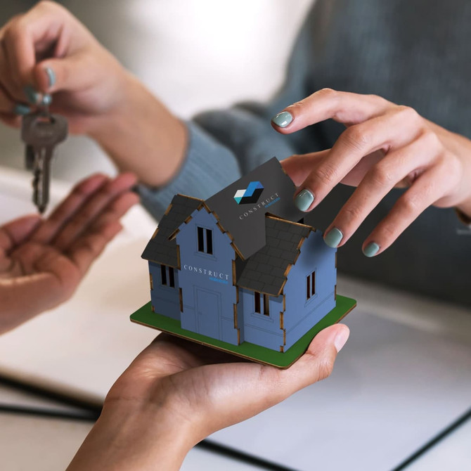 A small blue wooden model of a house with a green base and a logo on the roof, held in hands.