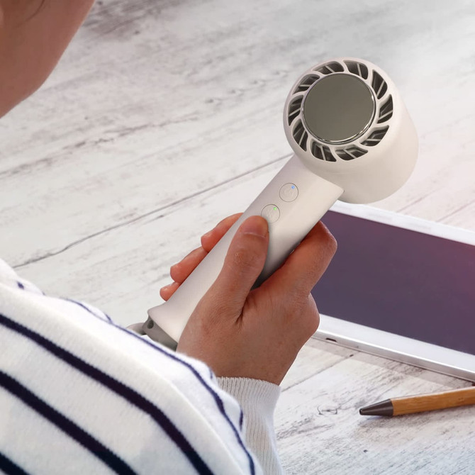 A handheld white cooling fan with a curved design, being held next to a tablet and a pen on a wooden surface.