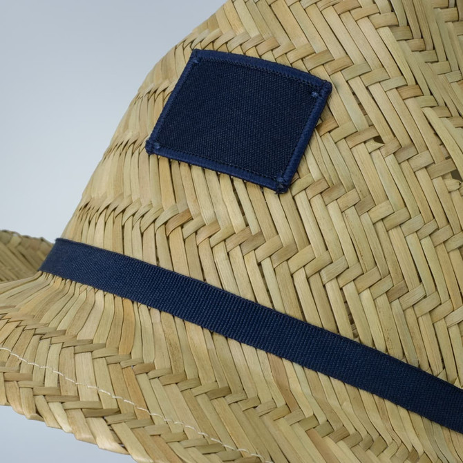 A straw fedora hat featuring a navy band and a square patch. The design showcases woven straw patterns.