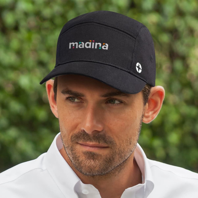 A black five-panel cap with a logo on the side, worn by a man against a greenery backdrop.