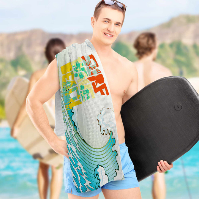 A sand-free towel in full colour featuring a wave design, held by a man at the beach with surfboards in the background.