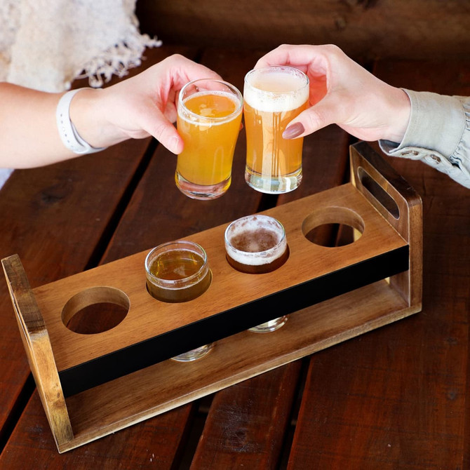 A wooden taster tray with five circular cutouts holding glasses of beer and small bowls. The tray has a rustic finish.