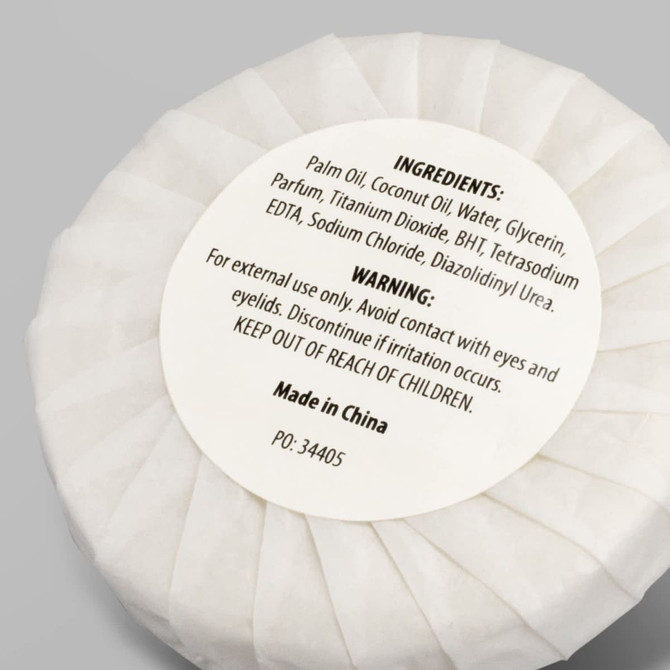 A white circular soap wrapped in paper, labelled with ingredients and warnings, features a logo.