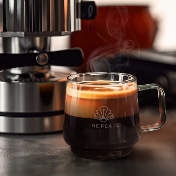 A coffee mug with a clear glass design, featuring layers of espresso and crema, placed near a coffee machine.
