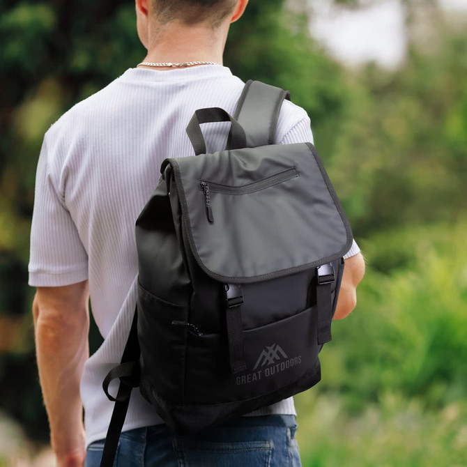 A black backpack with a flap and multiple compartments, carried by a person in outdoor surroundings.