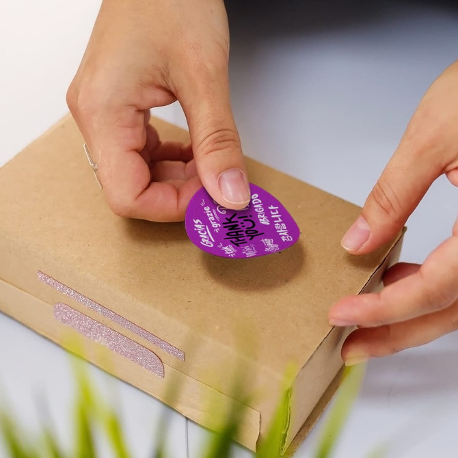A hand places a purple circular sticker on a brown cardboard package, featuring a logo and decorative elements.