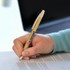 A kraft pen with a natural finish and a clip is held in a hand, writing on a paper document. The pen features a logo.