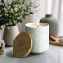 A white speckled candle with a bamboo lid, lit and surrounded by decorative home items and flowers.