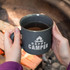 A matte enamel coffee mug in dark grey, held with two hands, filled with a dark beverage near a campfire.