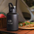 A drink bottle in matte black with a handle and silver cap, featuring a logo. Nearby are fruits, salad, and grilled food.