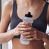 A drink bottle in clear plastic with a black lid, held by a person in a grey crop top, featuring a logo.