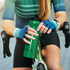 A green drink bottle with a black lid, held by a cyclist wearing blue gloves, sitting on a bicycle.