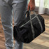 A black foldable duffle bag with grey handles being carried by a person in light pants. The bag features a zip closure.
