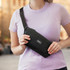 A black sling bag is held by a person wearing a light purple T-shirt, with a logo visible on the bag.