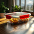 A clear lunch box with red clips contains bright yellow food items, set against a sunlit window.
