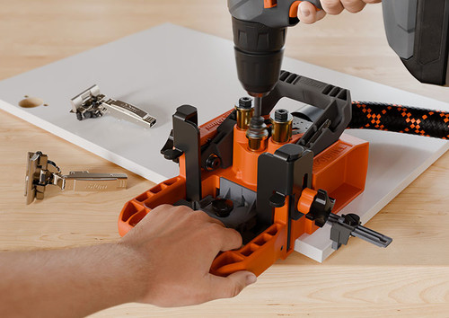 Blum ECODRILL jig used by Installer to drill hinge cup holes in a cabinet door. Quick-release clamp and integrated guides in use. Blum concealed hinges shown next to the workpiece.