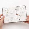 Hands holding a Making Memory Mini planner open to square-section notes filled with playlists, recipes, and stickers.