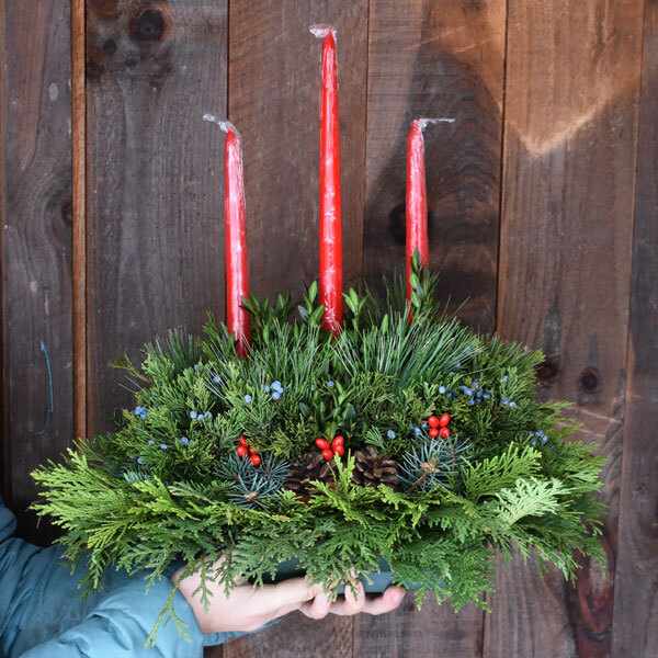 hands holding an evergreen centerpiece with three candles in front of a wood background