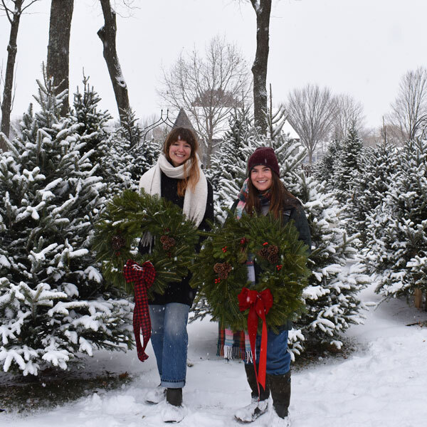 two women hold wreaths in front of outdoor christmas trees and snow