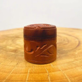Auric Blends Amber Butter Perfume - 1/5 oz. solid, small round container with an embossed design on the lid, placed on a wooden surface. Auric Blends Amber Butter Perfume - 1/5 oz. solid, small round container with an embossed design on the lid, placed on a wooden surface.