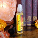 Shadow Scents Gold Perfume Oil - 1/3 oz. roll-on, a small clear bottle with a white cap and yellow label, placed on a wooden surface beside decorative salt lamps, books, and flowers.