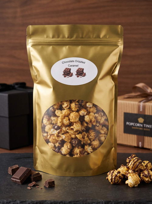 A flawless, perfectly smooth metallic gold stand-up pouch of scratch-made Chocolate Drizzled Caramel Popcorn. The precise label text 'Chocolate Drizzled Caramel' and the two distinct chocolate block graphics are visible on a sharp white circular sticker. A large clear window reveals the appetizing, detailed mix of caramel popcorn kernels heavily coated with rich dark chocolate drizzles. The bag is centered in a pristine, neutral professional studio environment, completely free of any popcorn tins