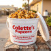 Colette's 1-gallon jumbo bucket filled with handcrafted copper-kettle caramel popcorn drizzled with rich dark chocolate, sitting outdoors along the Barefoot Landing boardwalk.
