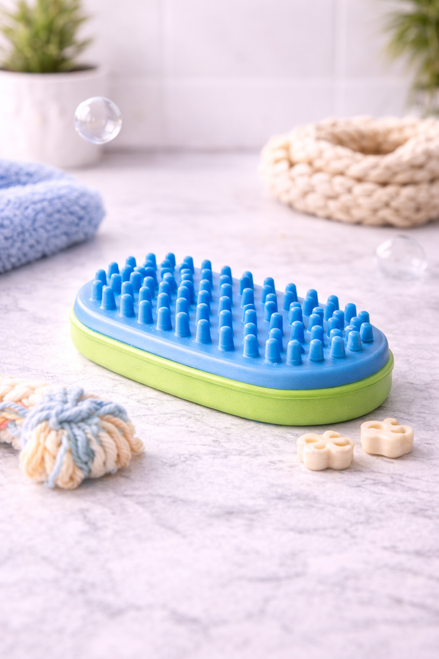 A pet grooming brush with soft rubber bristles and an ergonomic base, displayed on a clean countertop alongside pet toys and grooming accessories.