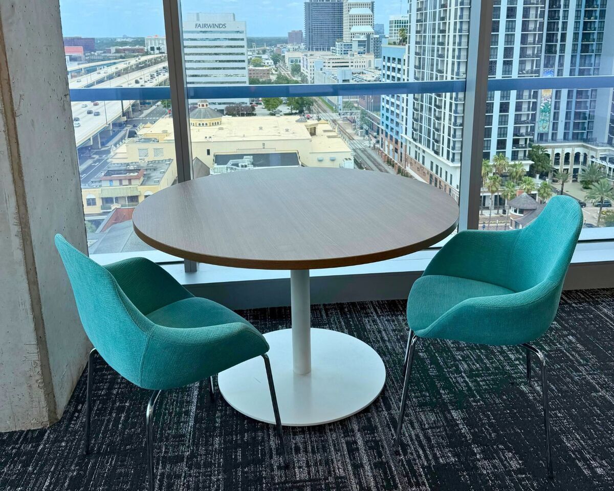 Round pedestal table with two teal upholstered chairs beside floor-to-ceiling windows