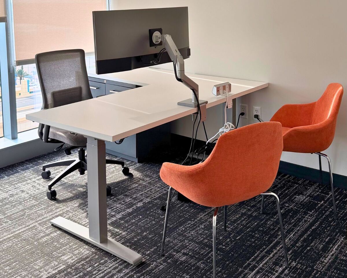 Private office with L-shaped sit-stand desk, monitor arm, task chair, and orange guest chairs