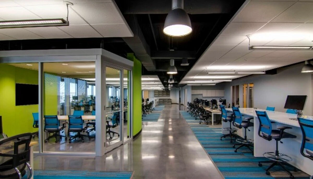 Open office aisle with white collaboration tables, blue drafting stools, and glass-enclosed meeting room