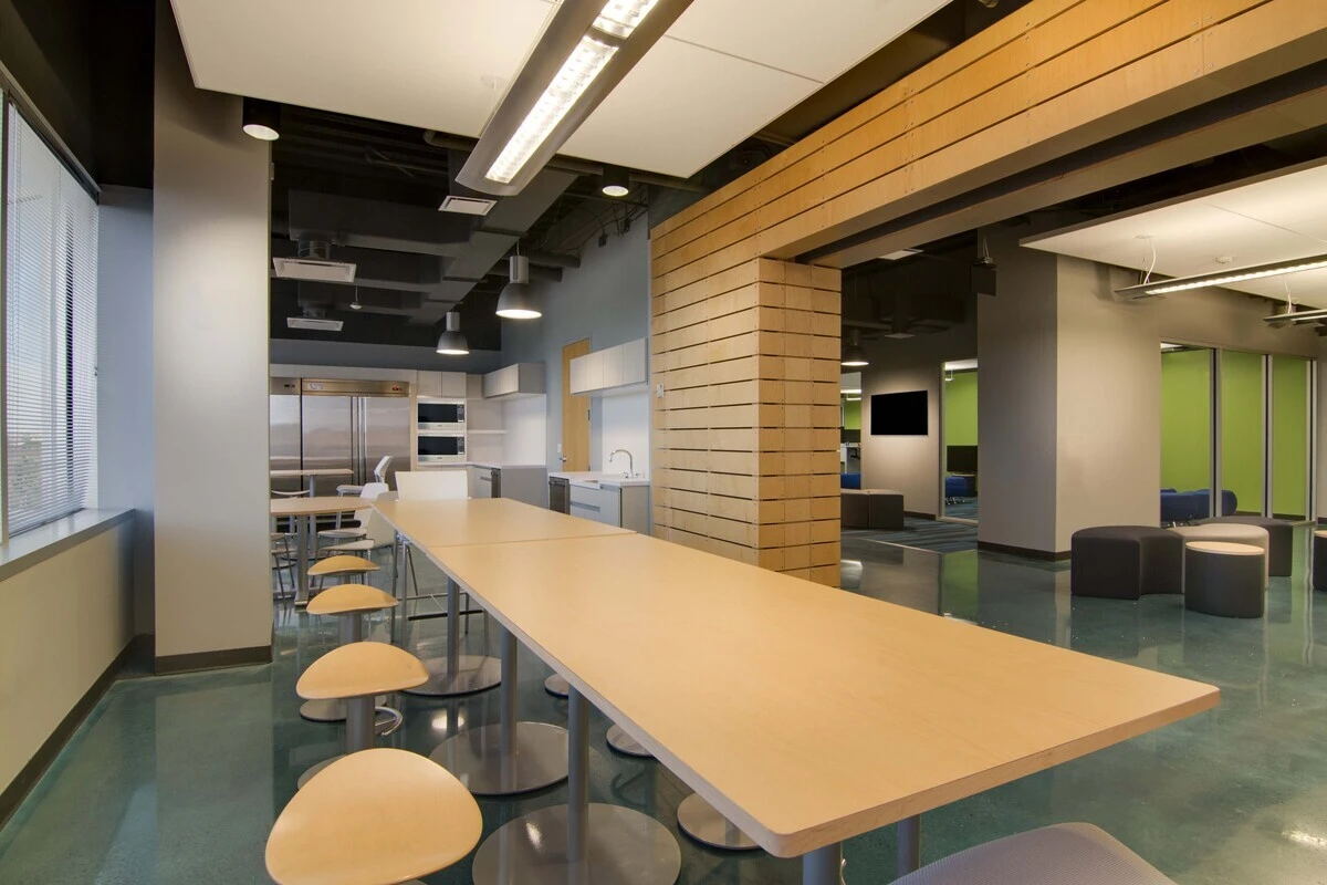 Break area with long maple high-top table, wood stools, and round ottoman seating